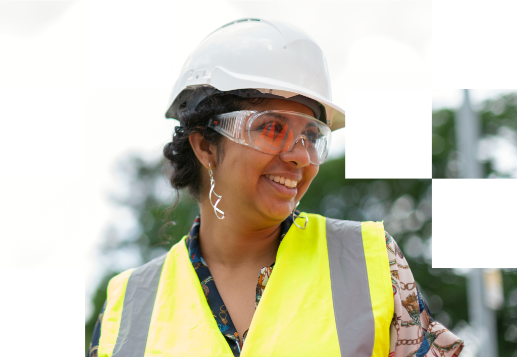 Woman wearing a hard hat and safety goggles, smiling while dressed in a high-visibility vest, representing the field service industry for independent contractors in elevator and HVAC sectors.